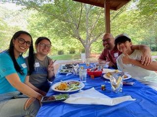 Four people seated at a picnic table with food, smiling under a pavilion.